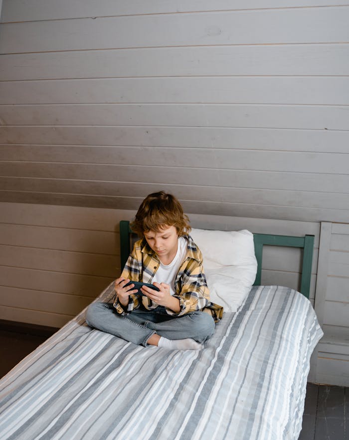 Young boy sitting on bed focused on mobile gaming indoors. Casual and relaxed atmosphere.